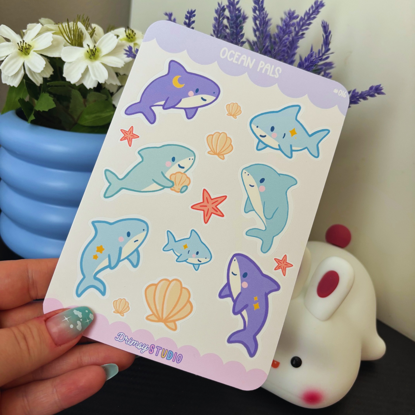 A hand holds a sticker sheet featuring cute sharks, shells, and starfish, with flowers in the background.