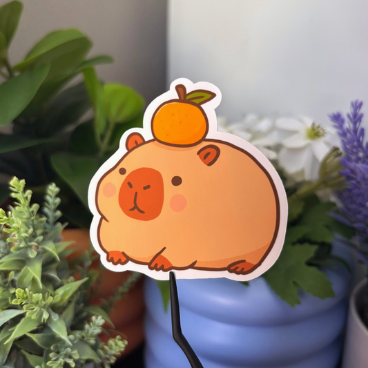 Orange capybara sticker with an orange on its head, held by black tweezers.